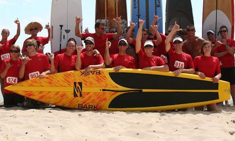 Participants in a race held by Surftech pose behind one of Surftech's many high-performing stand up paddle boards. Photo Credit: A Day At the Beach